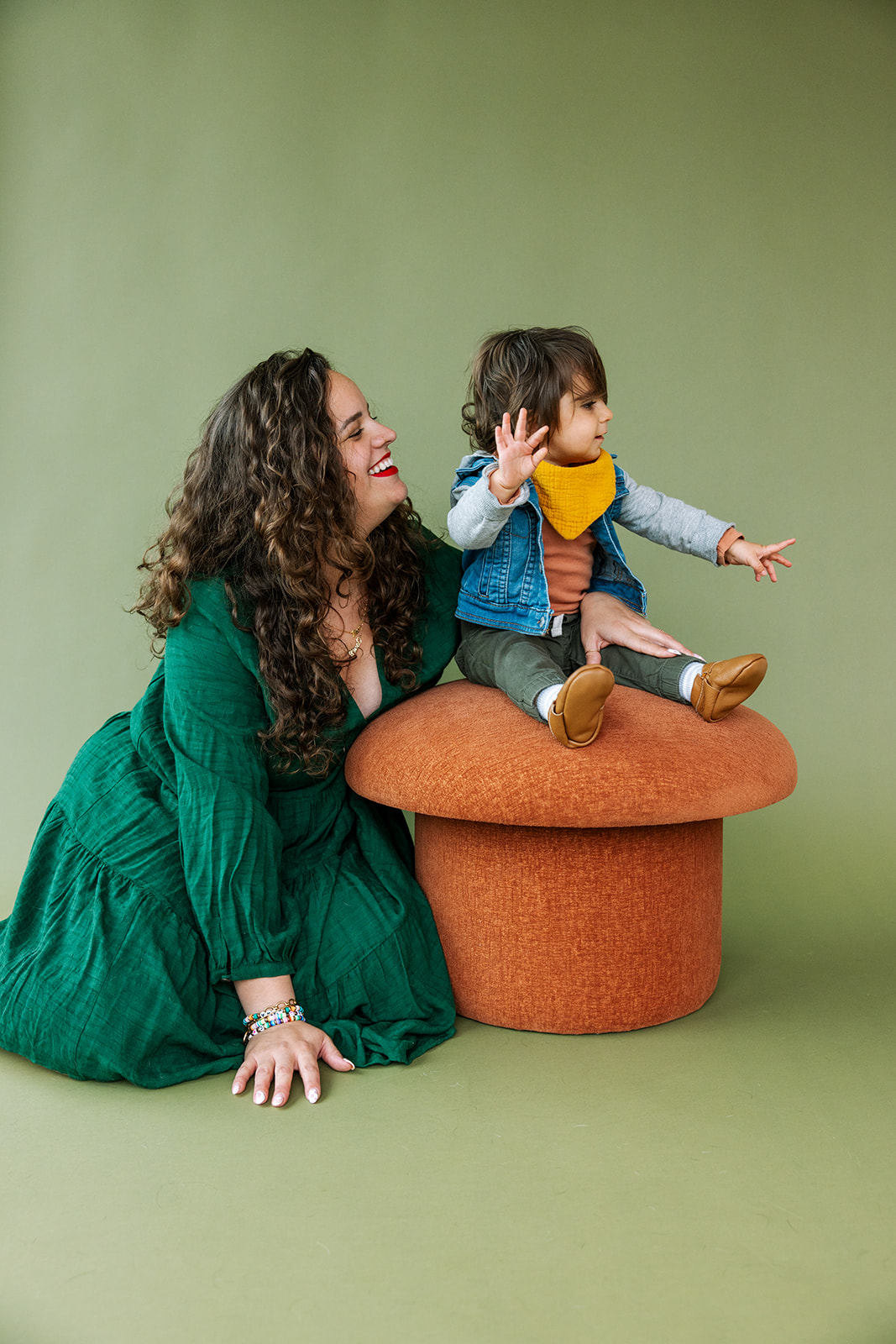 photo of mom in green dress sitting on ground next to toddler sitting on an orange mushroom shaped stool