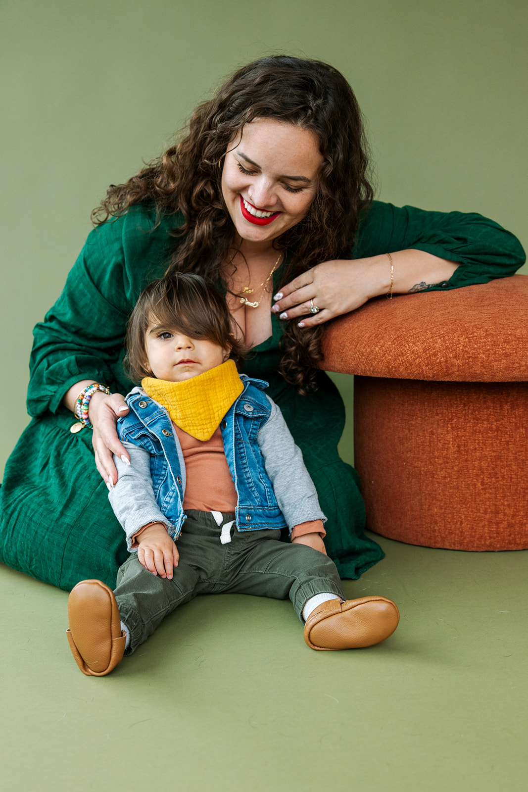 photo of mom sitting on green paper backdrop behind toddler son and leaning on an orange mushroom shaped stool