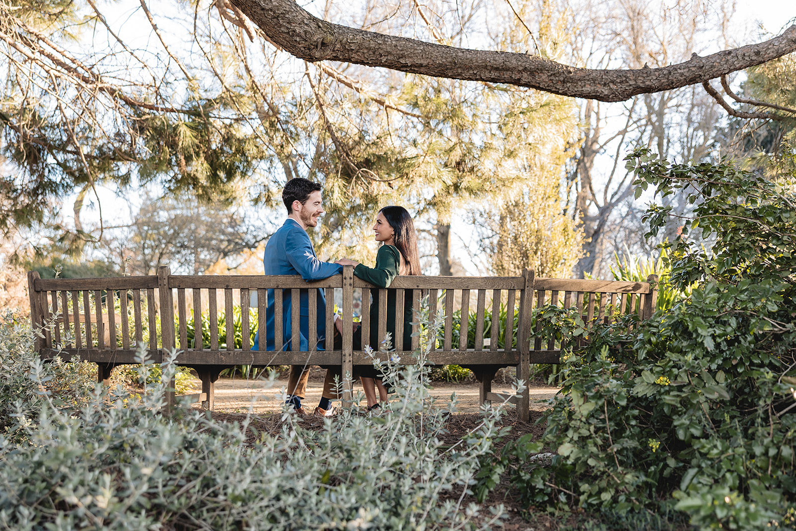 Couple sites on a bench and looks at each other lovingly surrounded by greenery. Photo by sacramento photographer.