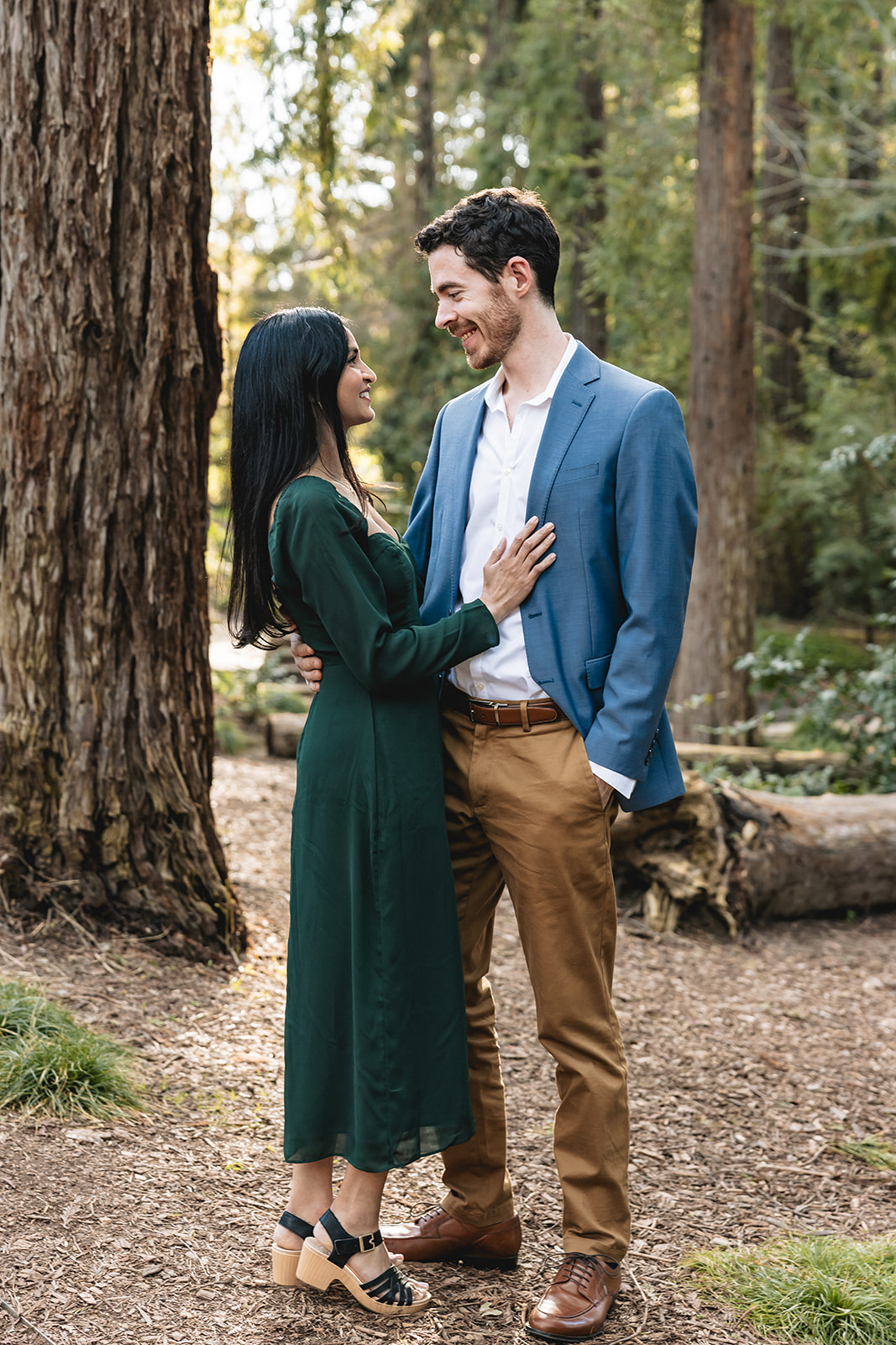 Engaged couple looks at each other smiling in the middle of the woods. 