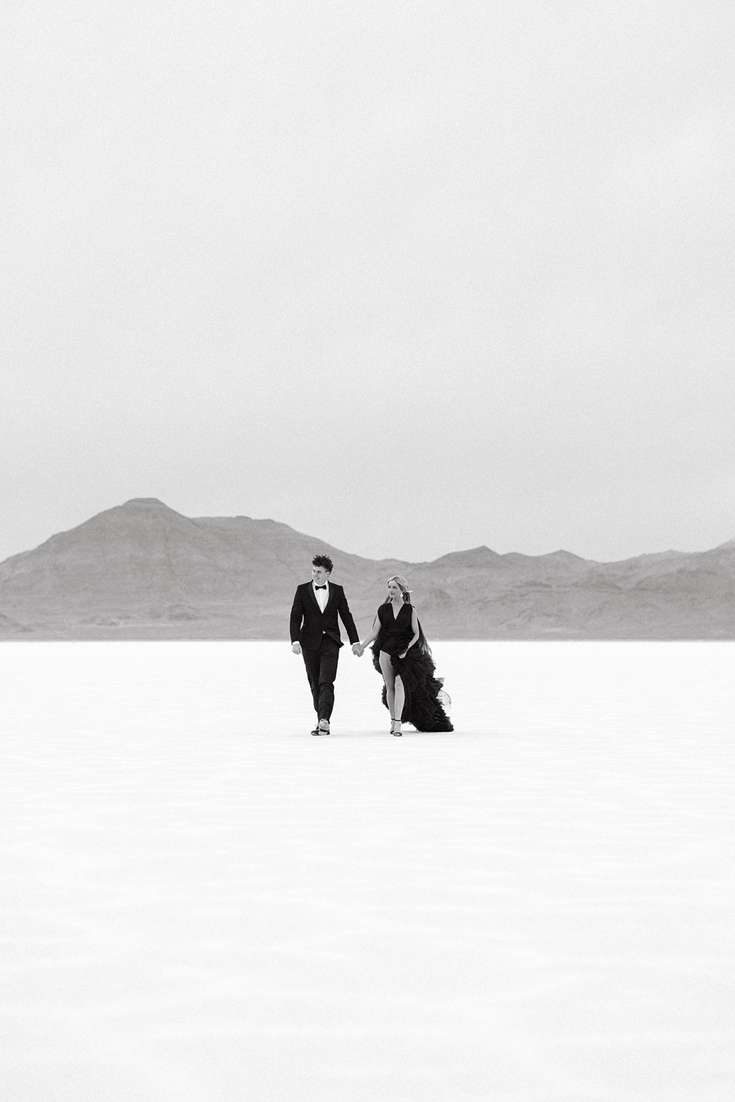 black and white photo of couple wearing formal attire walking forward with the salt flats mountains in the backgroud