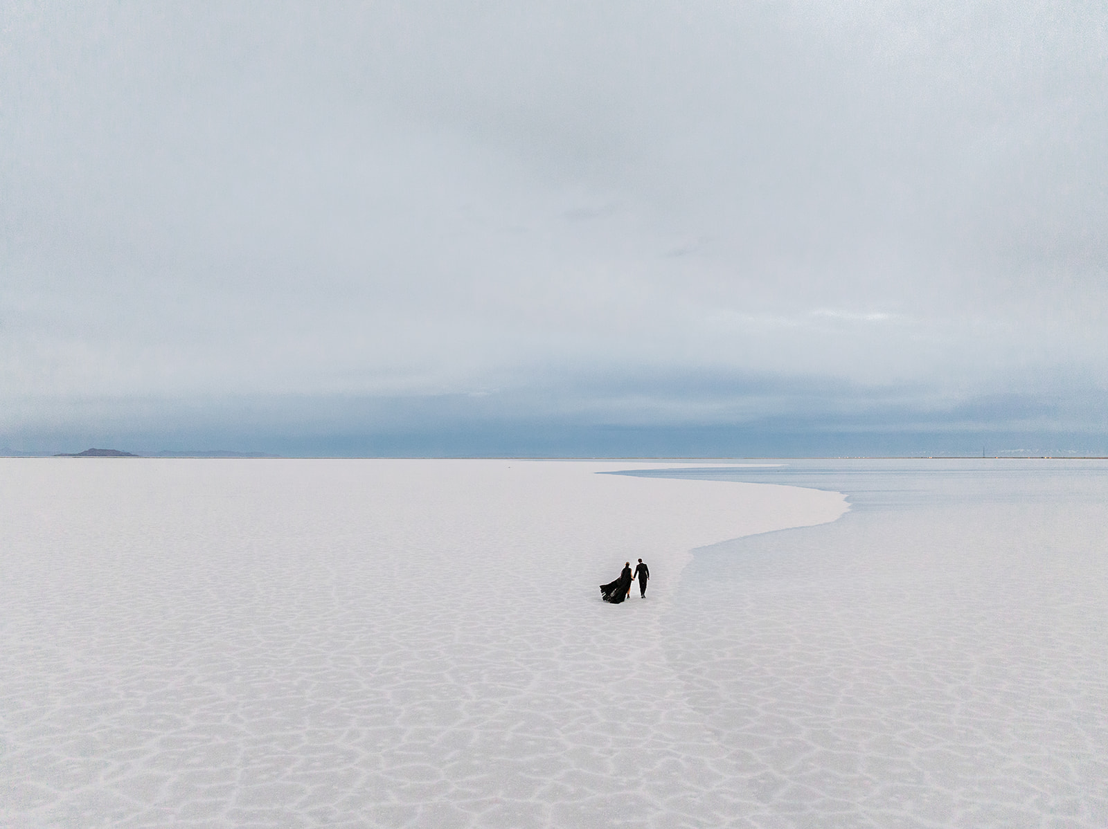 Utah Salt Flats engagement drone photo with dramatic black gown flowing while the couple is holding hands and walking